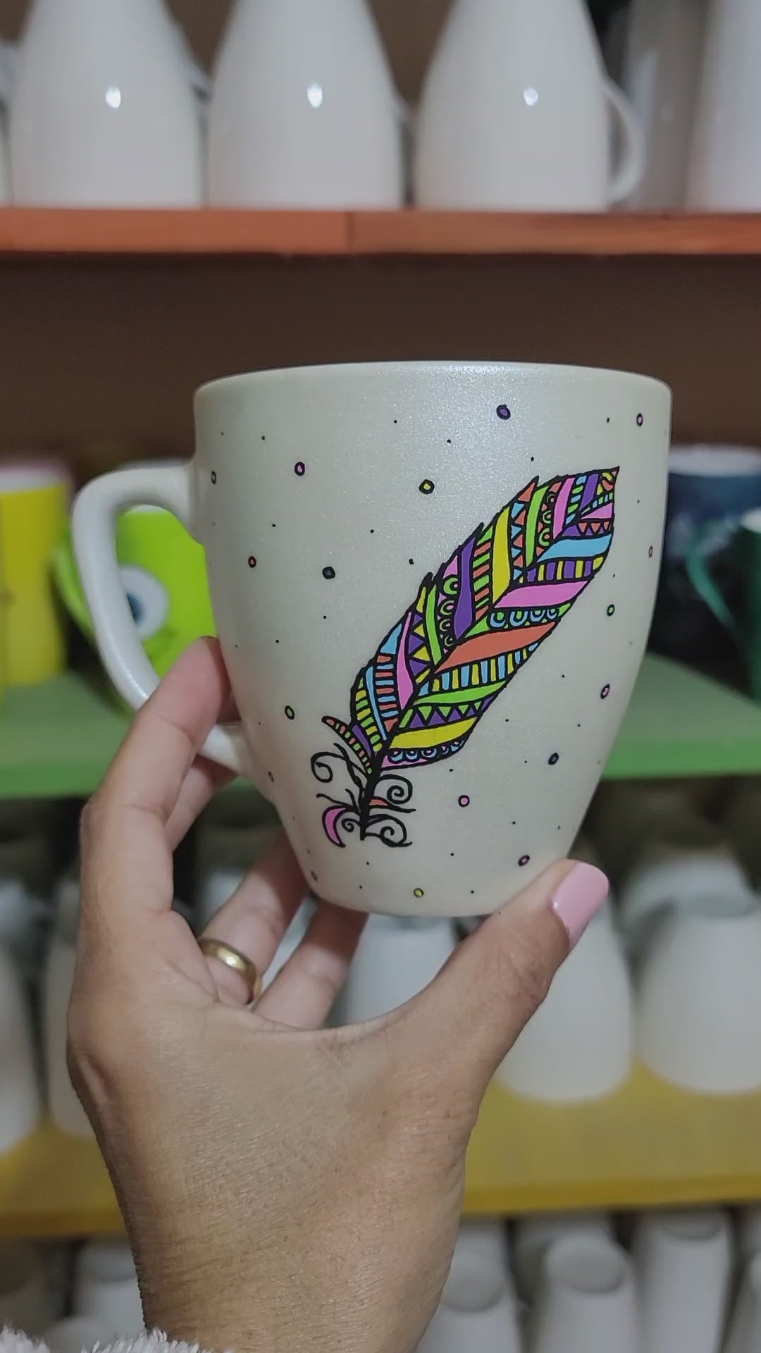 A Pearl white hand painted mug with a colorful feather outlined in black and filled with paterns in colors of orange, yellow, pink, purple, green and blue and the name "Emma" written on the other side