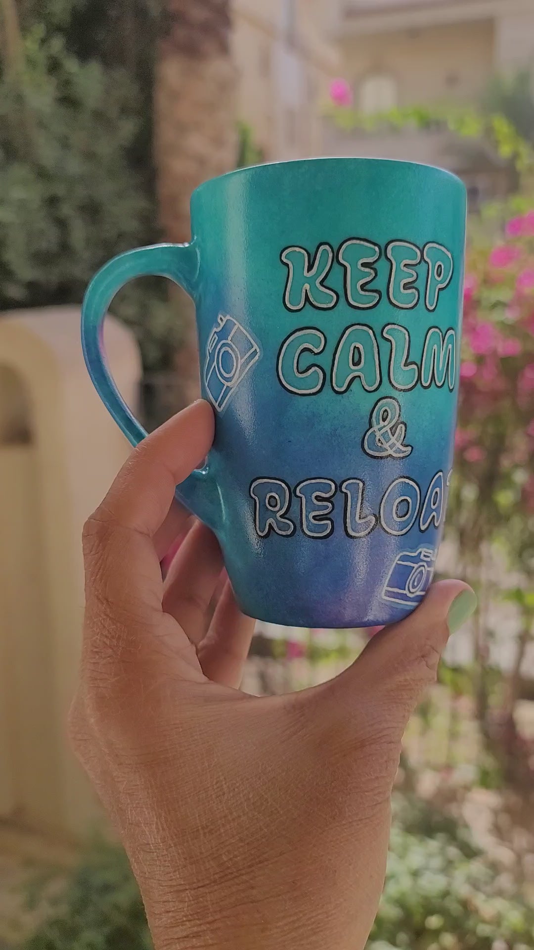 A Blue hand painted mug with the words "Keep Calm and reload" and small cameras scattered all over the mug and the name Aly on the other side