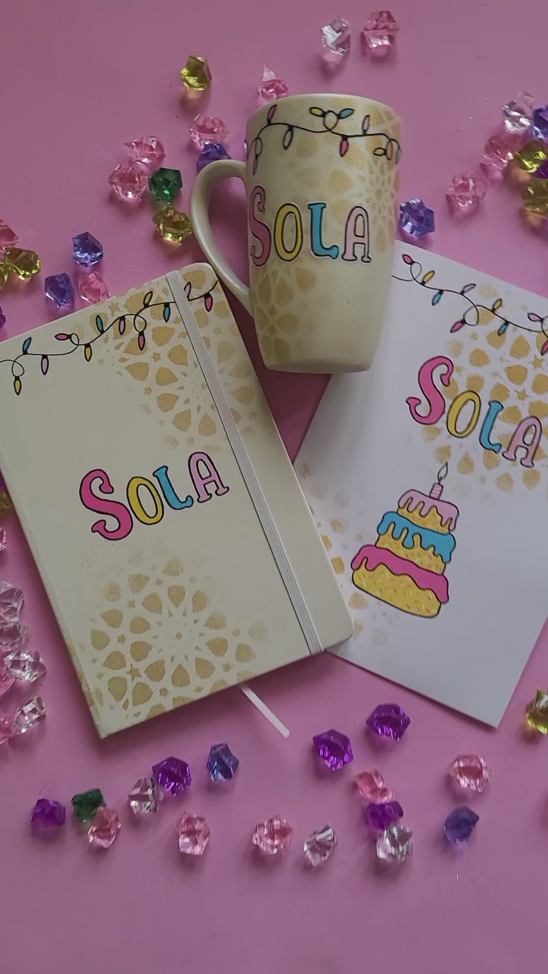 A Gift Set of A matching mug, notebook and card . All handpainted in ivory with geometric patterns in gold, and have festive lights all over the top. The notebook has the words "Strong Independent Cutest Woman" the mug has a cake design and the card has the name sola and a cake design on it