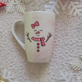 A Pearl white hand painted mug with a red bow drawn on the side and 2 closed eyes and an orange nose and a mouth smiling. wearing a red scarf and 3 buttons drawn below it and 2 tree branches as hands and the name 