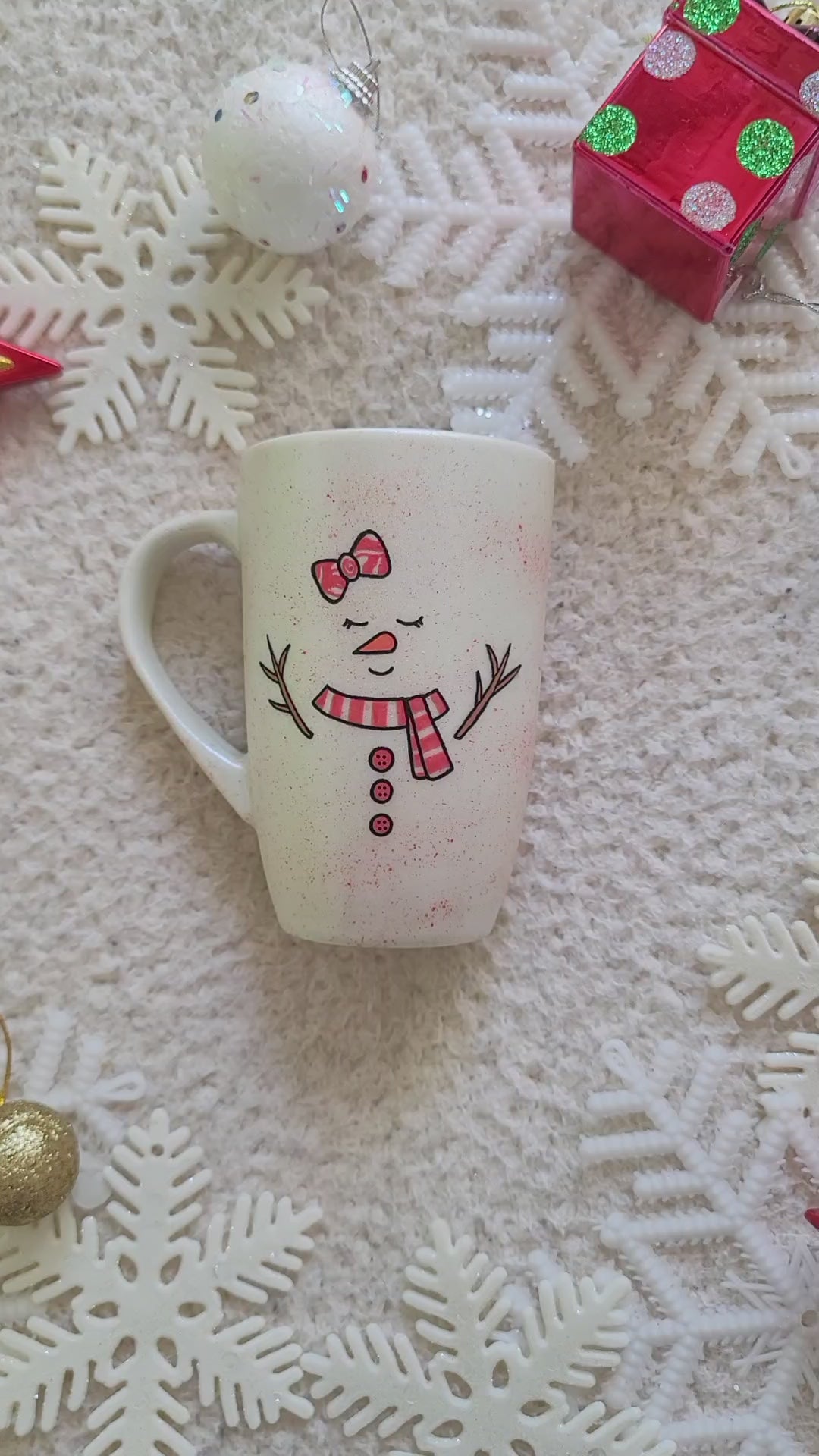 A Pearl white hand painted mug with a red bow drawn on the side and 2 closed eyes and an orange nose and a mouth smiling. wearing a red scarf and 3 buttons drawn below it and 2 tree branches as hands and the name "Emma" on the other side