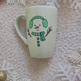 A pearl white hand painted mug with a snow boy painted wearing a green Earmuffs and a green scarf with 3 green buttons in the center and hands made out of tree branches. with an orange nose and a smiling mouth. and the other side has the name 