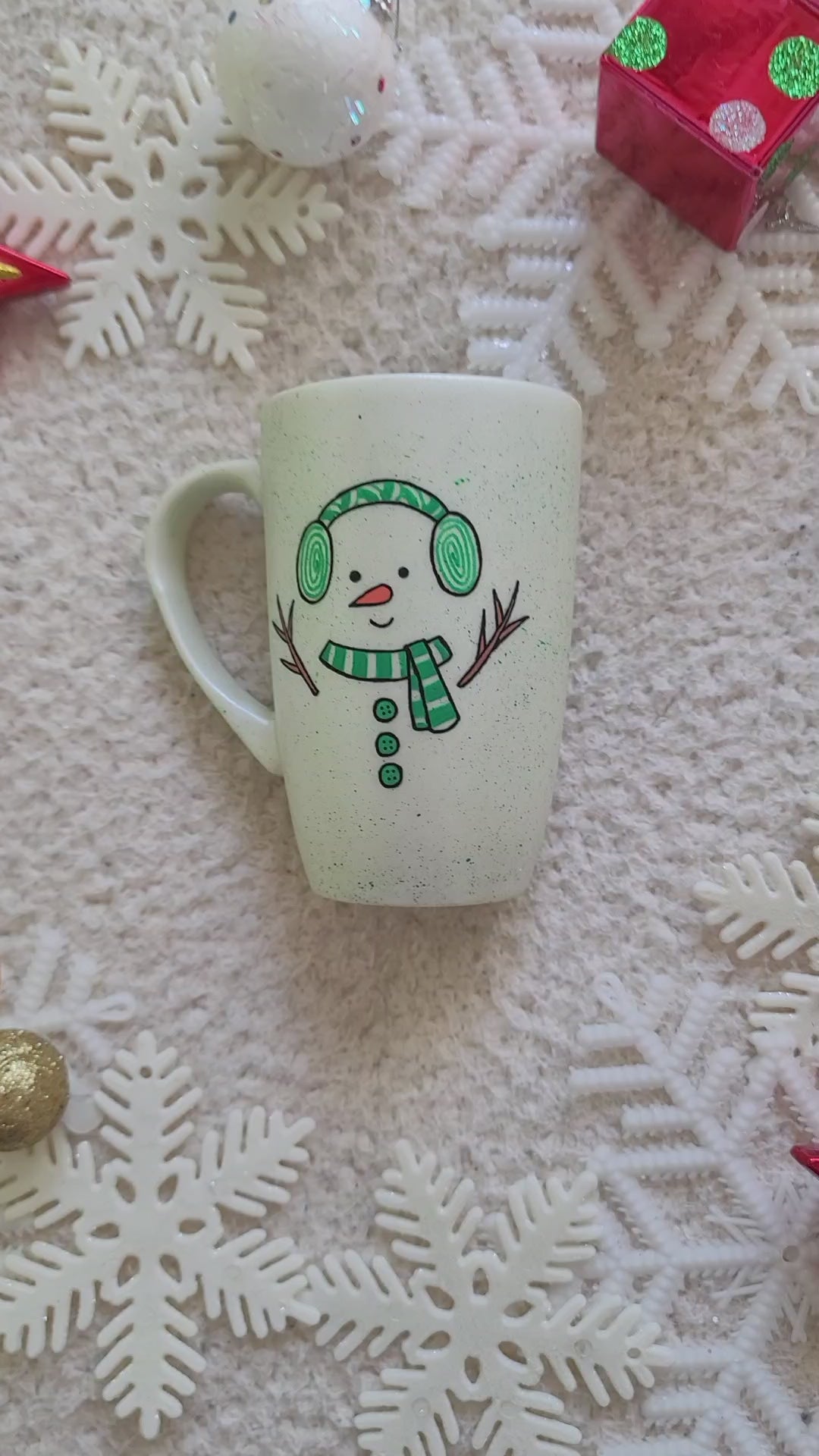 A pearl white hand painted mug with a snow boy painted wearing a green Earmuffs and a green scarf with 3 green buttons in the center and hands made out of tree branches. with an orange nose and a smiling mouth. and the other side has the name "Hamza"