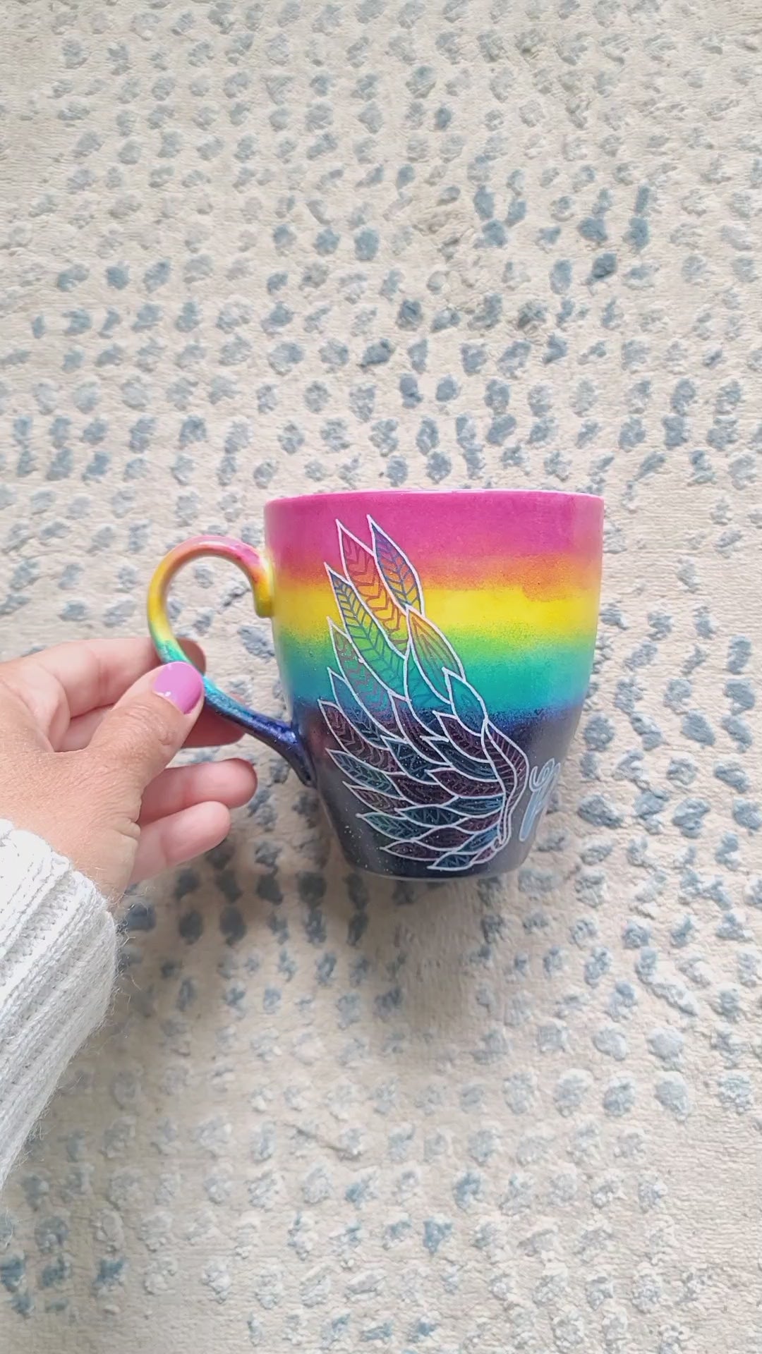 A gradient hand painted mug(rainbow colors in the top half and black galaxy in the bottom" with a wing drawn on one side and the name "poussy" in the center and another wing on the other side