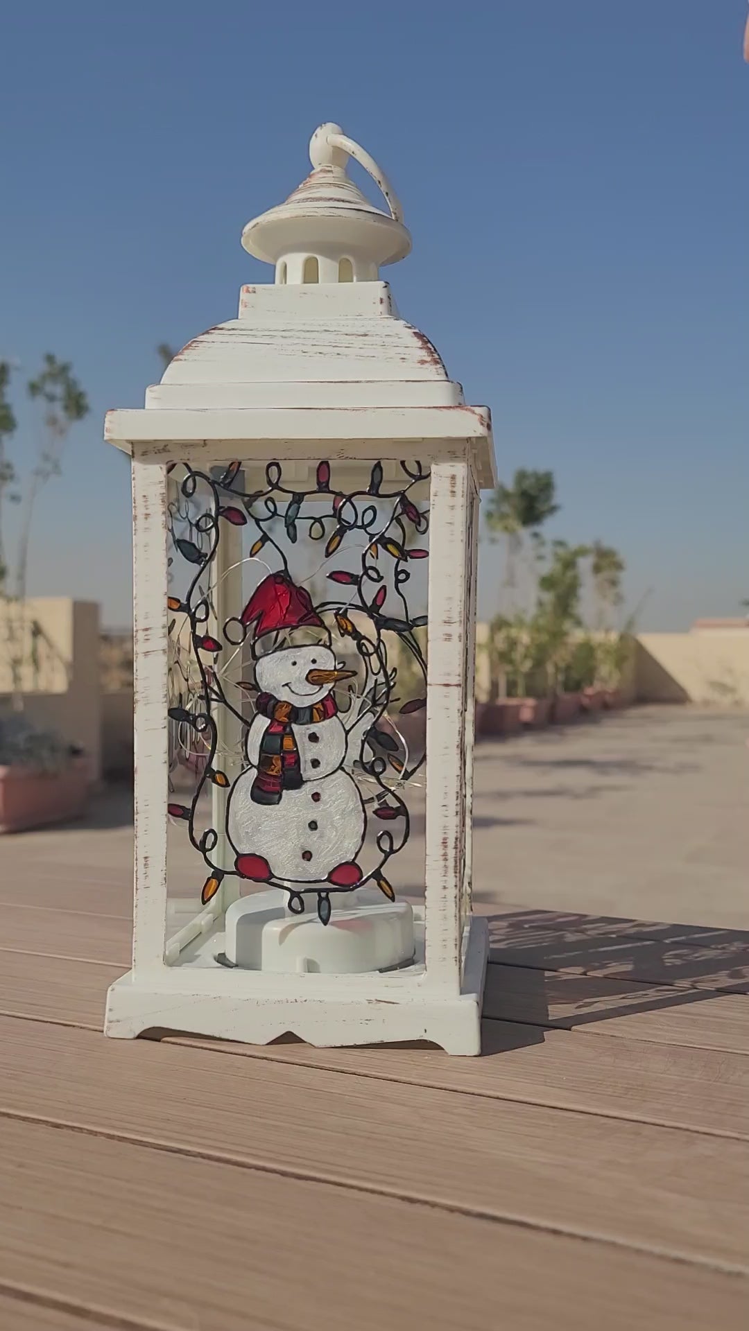 A White Vintage Lantern with 4 hand painted glass sides.  the first with a snowman, the second with the name "Emma" the third with a reindeer, and the fourth with the letter E in patterns of red white and green. And all four glasses have festive lights painted on the top part