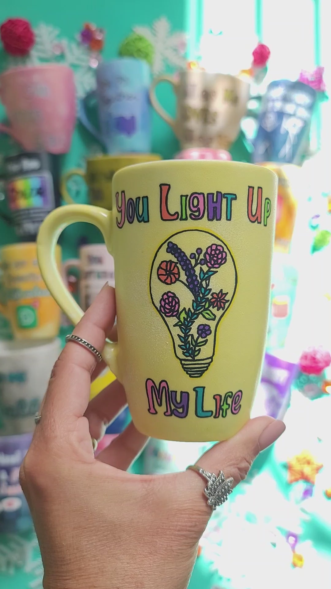 A Pastel yellow hand-painted mug with the words "you light up my life" and a bulb drawn on it with flowers inside. and the other side has the name "menna"