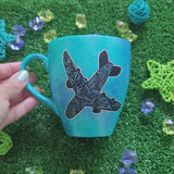 A Blue hand apinted mug with an airplane painted on it full of patterns in grey and the other side has the words 