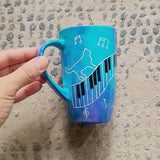 A Blue Hand painted mug with piano keys drawn moving all over hz on the mug and cats on it and below it , with musical symbols scattered and the letter M on the other side