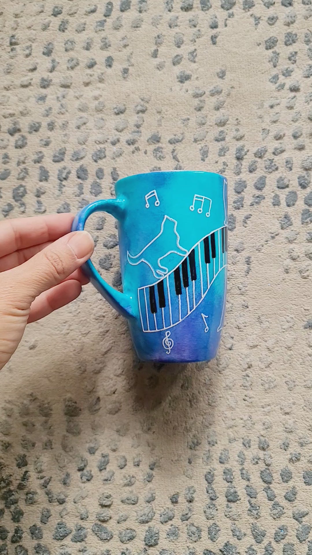 A Blue Hand painted mug with piano keys drawn moving all over hz on the mug and cats on it and below it , with musical symbols scattered and the letter M on the other side