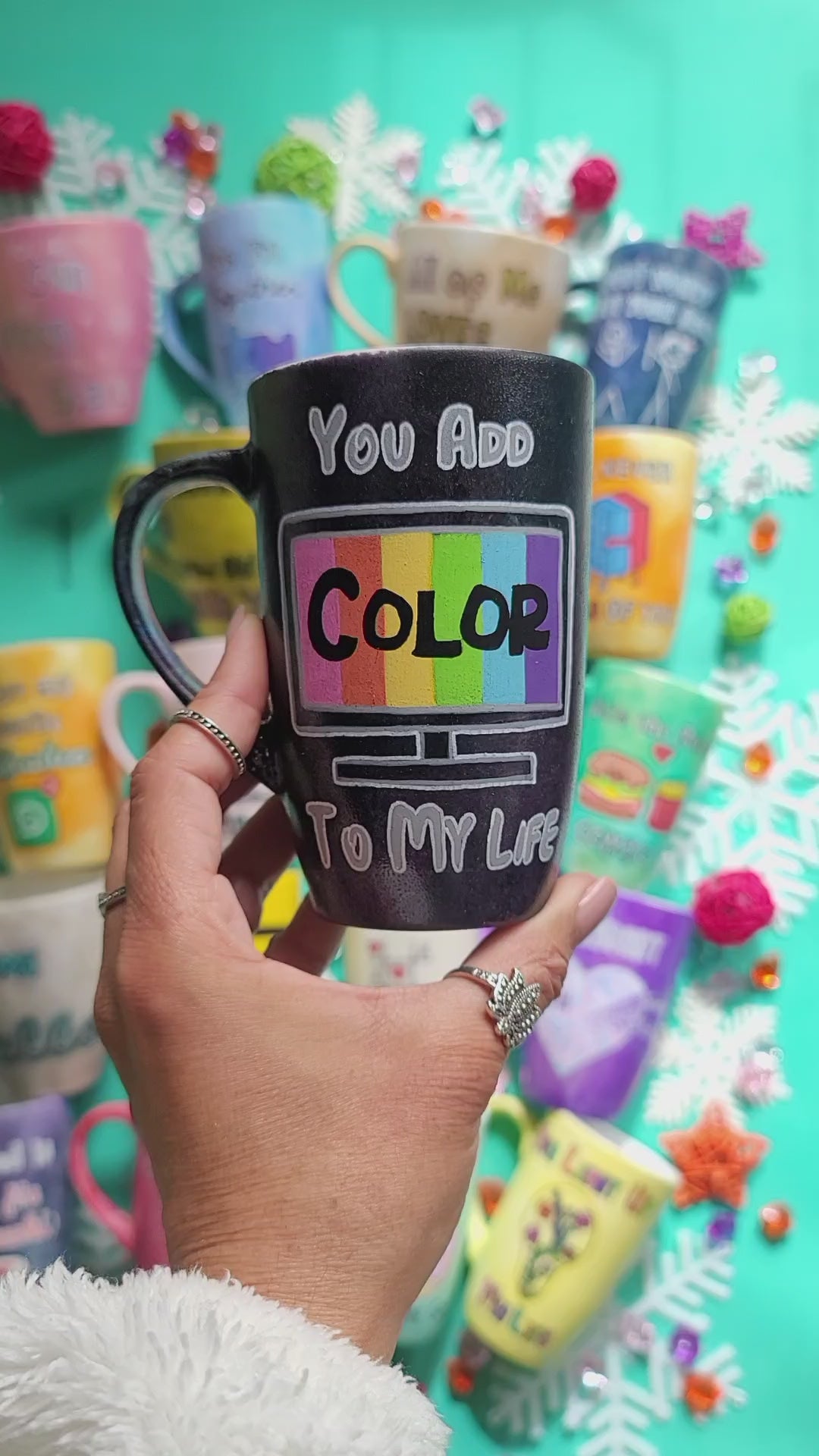 A hand-painted black mug with the words "you add" on the top center, below it a tv led screen drawn with colors inside and the word "color" in black. then below it the words " to my life".on the other side of the mug the name "Nourhan"