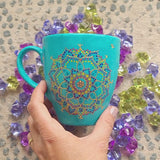 A hand painted blue Indian color mug with a mandala painted in gold and on the other side the words 