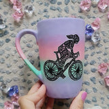 A gradient hand painted mug (lilac-pink-mint-lilac) with a silhouette of a girl on a bike drawn and filled with patterns and the other side has the name Bambie