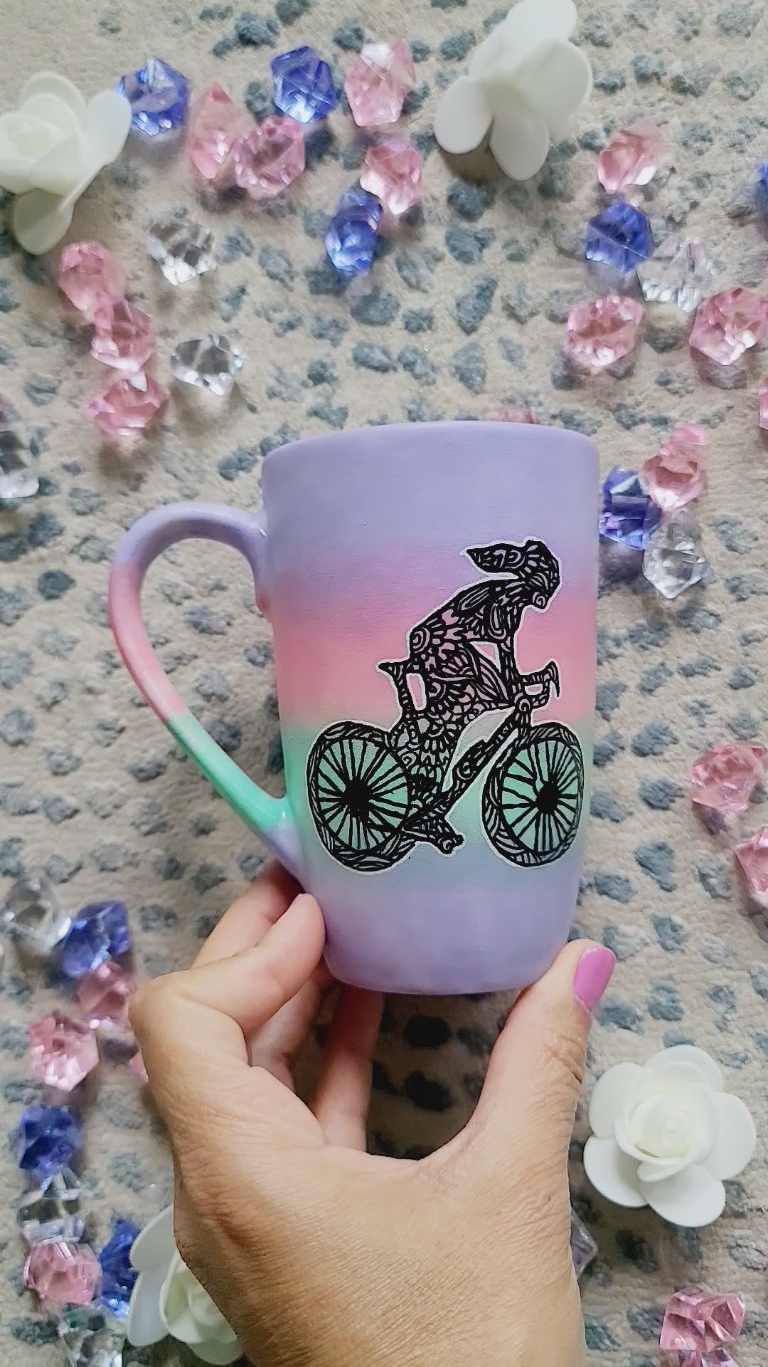 A gradient hand painted mug (lilac-pink-mint-lilac) with a silhouette of a girl on a bike drawn and filled with patterns and the other side has the name Bambie