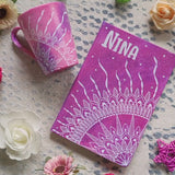 A Gift set of a matching mug and notebook both hand painted in pink galaxy color and have a corner mandala drawn in white. Both have the name Nina written on them