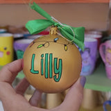 A gold hand painted ball ornament with festive lights all around the top and the name 