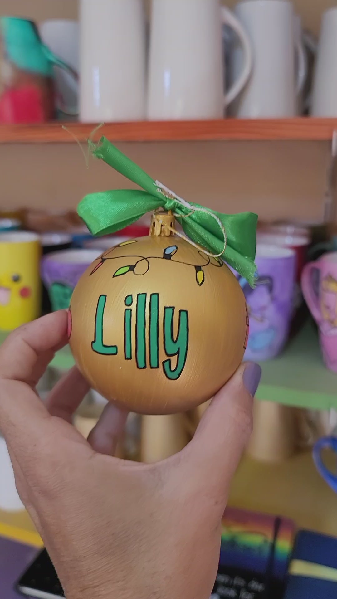 A gold hand painted ball ornament with festive lights all around the top and the name "Lilly" written in green outlined with black