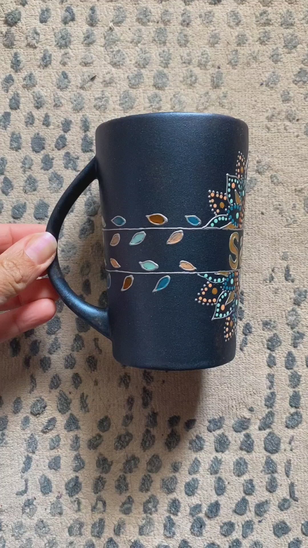 A Shimmery blue hand painted mug with a mandala split into 2 with a space in between that has the name "Salma" and the mandala is outlined with silver relief and dot art of gold, turquoise, bronze and bej