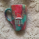 A Christmas galaxy hand painted mug with a reindeer antlers with a santa hat and reindeer ears, then below it the name 