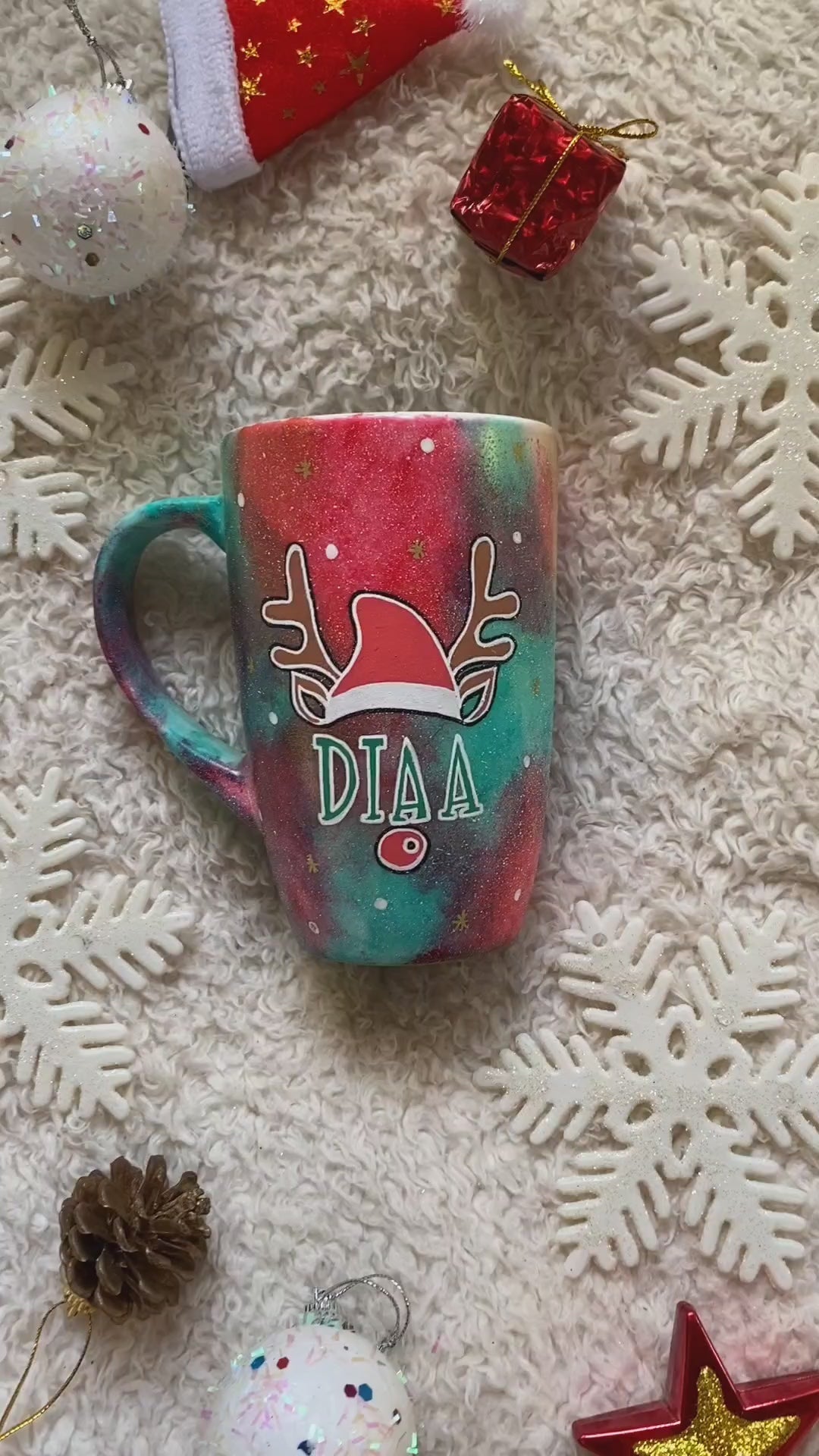 A Christmas galaxy hand painted mug with a reindeer antlers with a santa hat and reindeer ears, then below it the name "Diaa" and below it the reindeer nose, with white dots and gold stars scattered all over the mug and a tree drawn in white on the other side with a gold star on top