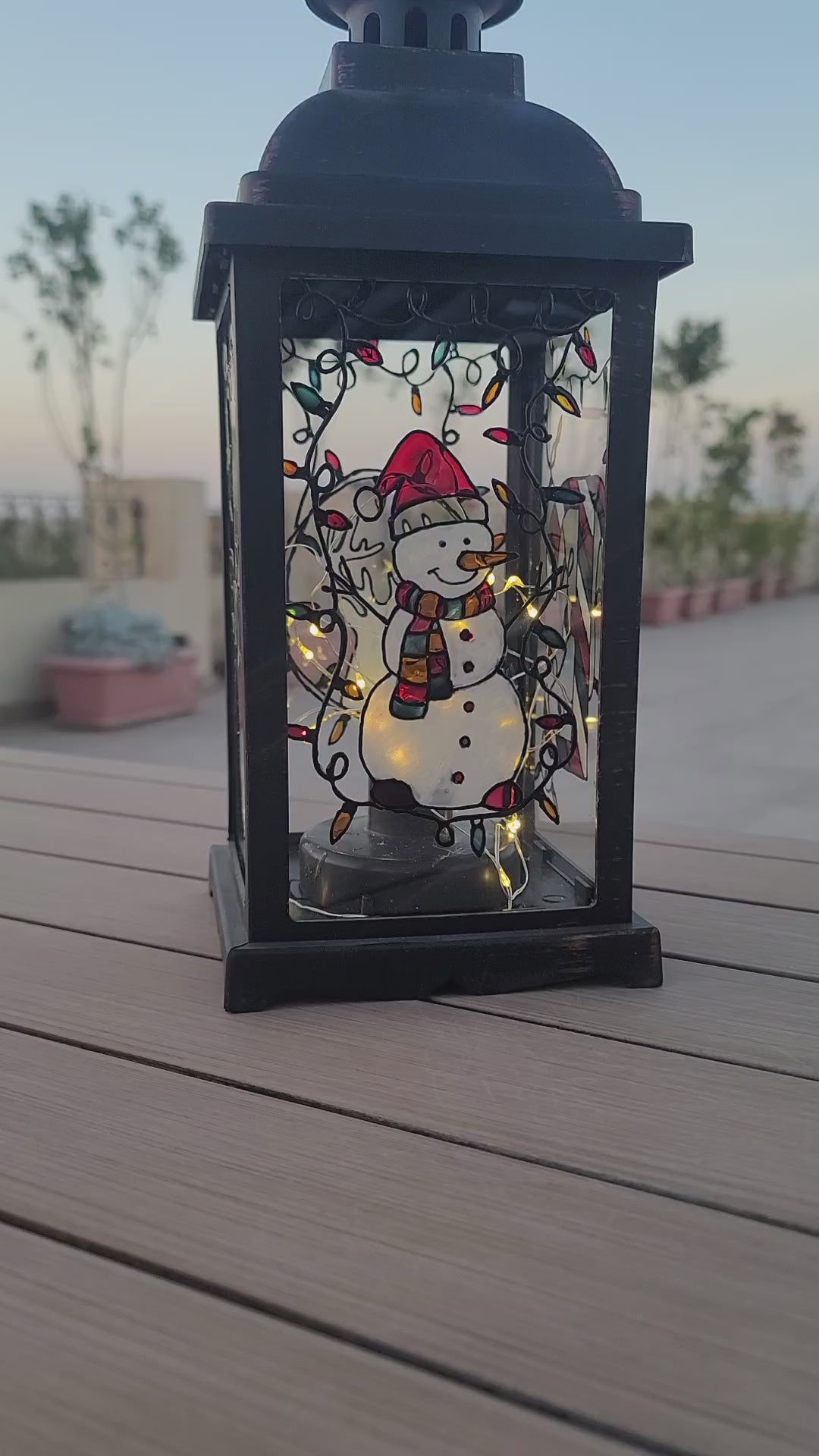 A Black Vintage Lantern with 4 hand painted glass sides.  the first with a snowman, the second with the name "Emma" the third with a reindeer, and the fourth with the letter E in patterns of red white and green. And all four glasses have festive lights painted on the top part