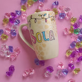 A Pearl white hand painted mug with a layered cake painted  in yellow and icing of baby pink , blue and fushia, with sprinkles all over the yellow cake and a pink candle on top, also the mug has birthday lighting all over the top of the mug. and the name 