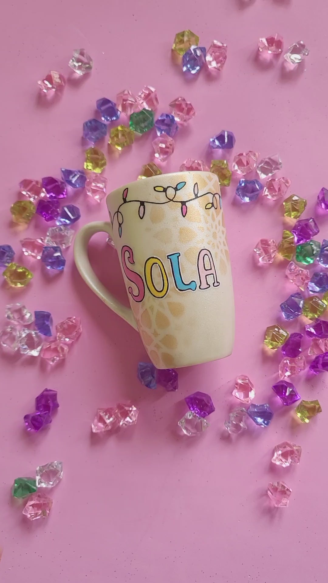 A Pearl white hand painted mug with a layered cake painted  in yellow and icing of baby pink , blue and fushia, with sprinkles all over the yellow cake and a pink candle on top, also the mug has birthday lighting all over the top of the mug. and the name "Sola" on the other side