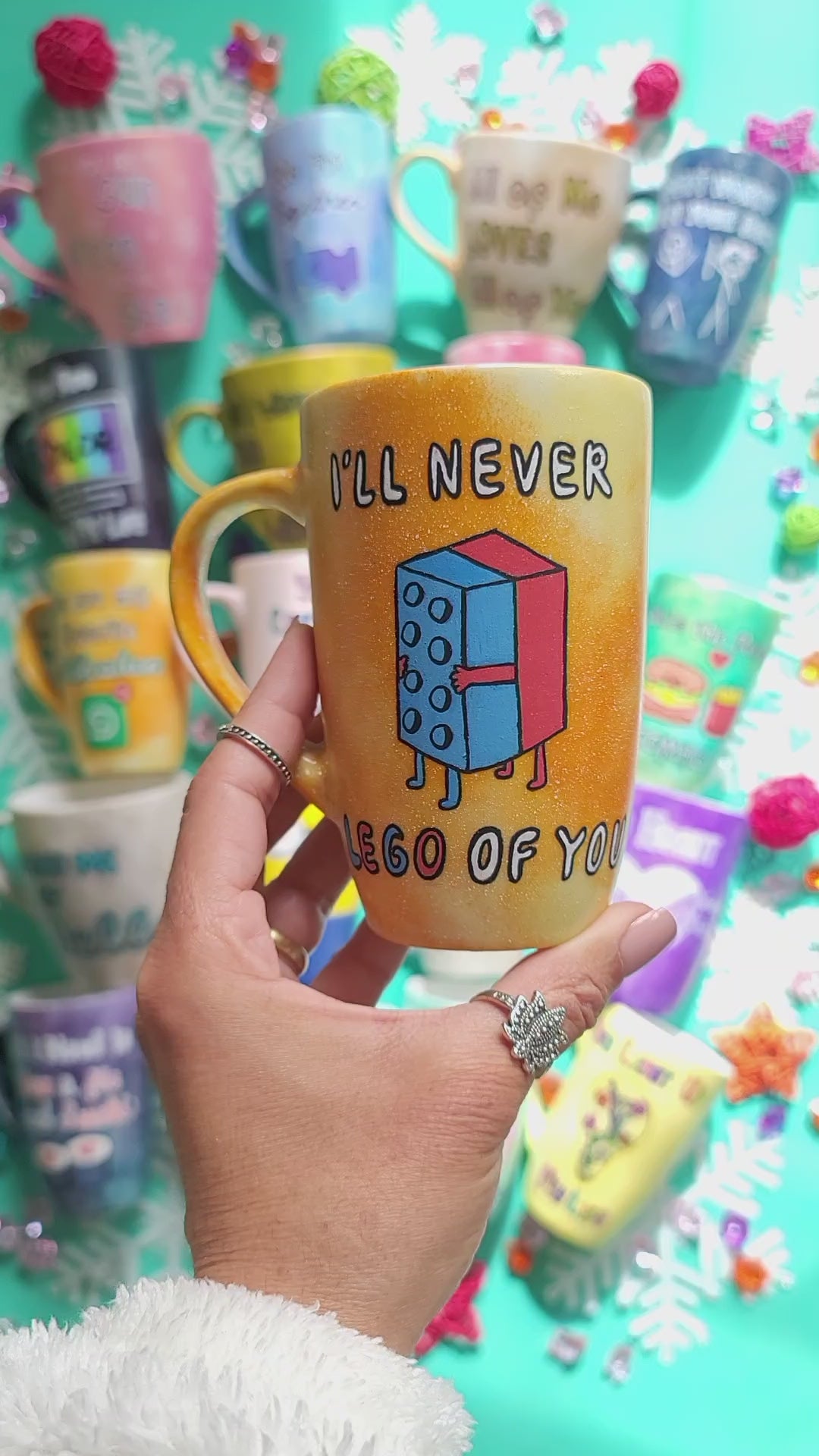 A yellow orange hand-painted mug with the words "I'll never" and below it two lego pieces hugging each other and the below it the words "LEGO of you" and on the other side the name "Reham"