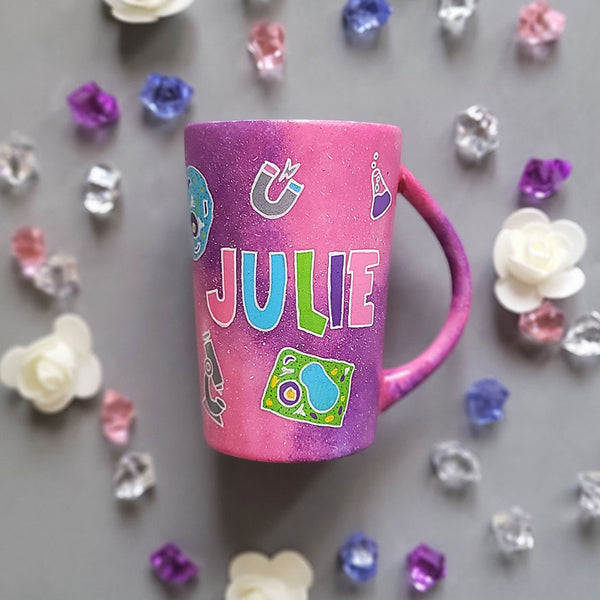 A Pink Galaxy hand painted mug with the name Julie written in the center and science items scattered al over "plant cell, animal cell, telescope, magnet....etc."