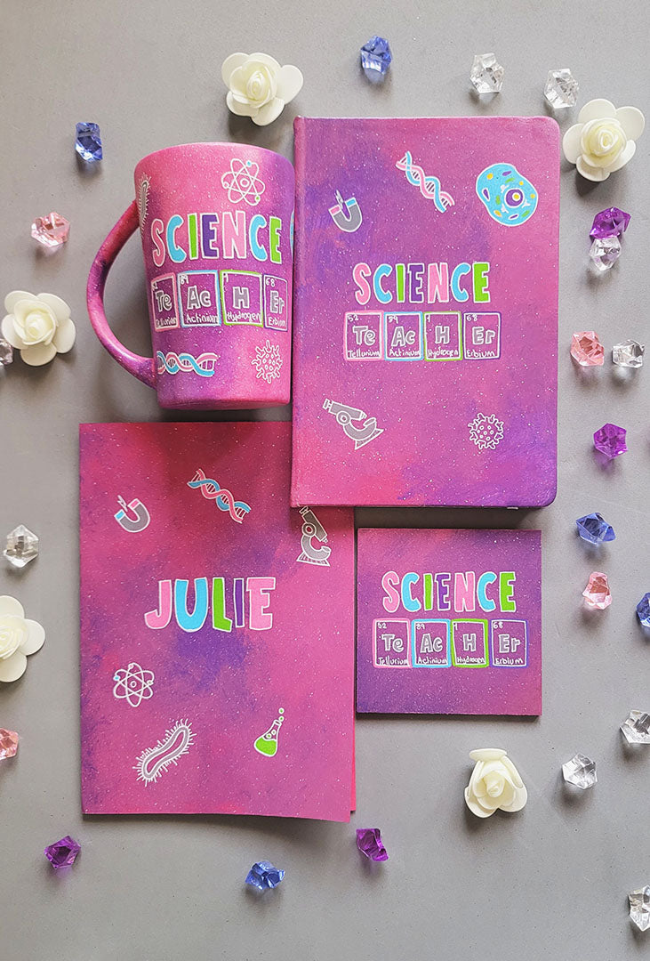 A Hand painted Gift set containing Mug, Notebook, Card and Coaster all in pink galaxy color with the word Science on it and under it the word teacher written in the form of periodic table elements. with science items scattered all over. and the other side of the mug, notebook and card all have the name Julie