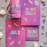 A Hand painted Gift set containing Mug, Notebook, Card and Coaster all in pink galaxy color with the word Science on it and under it the word teacher written in the form of periodic table elements. with science items scattered all over. and the other side of the mug, notebook and card all have the name Julie