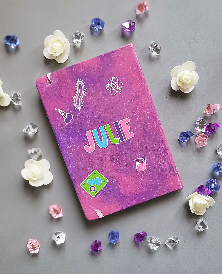 A Pink Galaxy hand painted Notebook with the name Julie written in the center and science items scattered al over "plant cell, animal cell, telescope, magnet....etc."