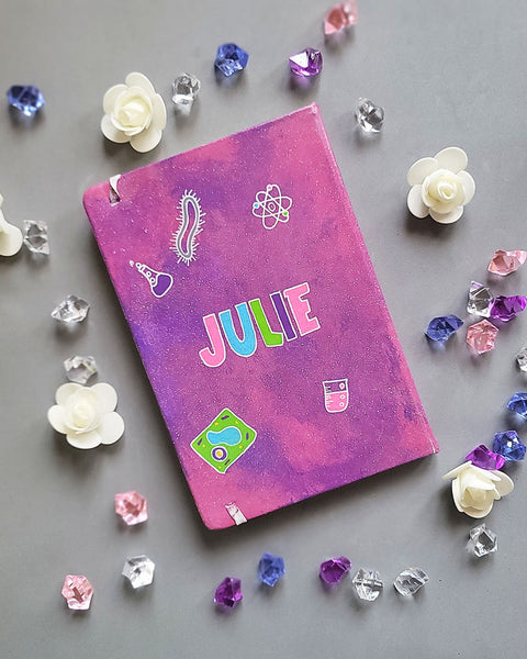 A Pink Galaxy hand painted Notebook with the name Julie written in the center and science items scattered al over "plant cell, animal cell, telescope, magnet....etc."