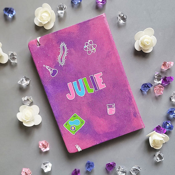 A Pink Galaxy hand painted Notebook with the name Julie written in the center and science items scattered al over "plant cell, animal cell, telescope, magnet....etc."