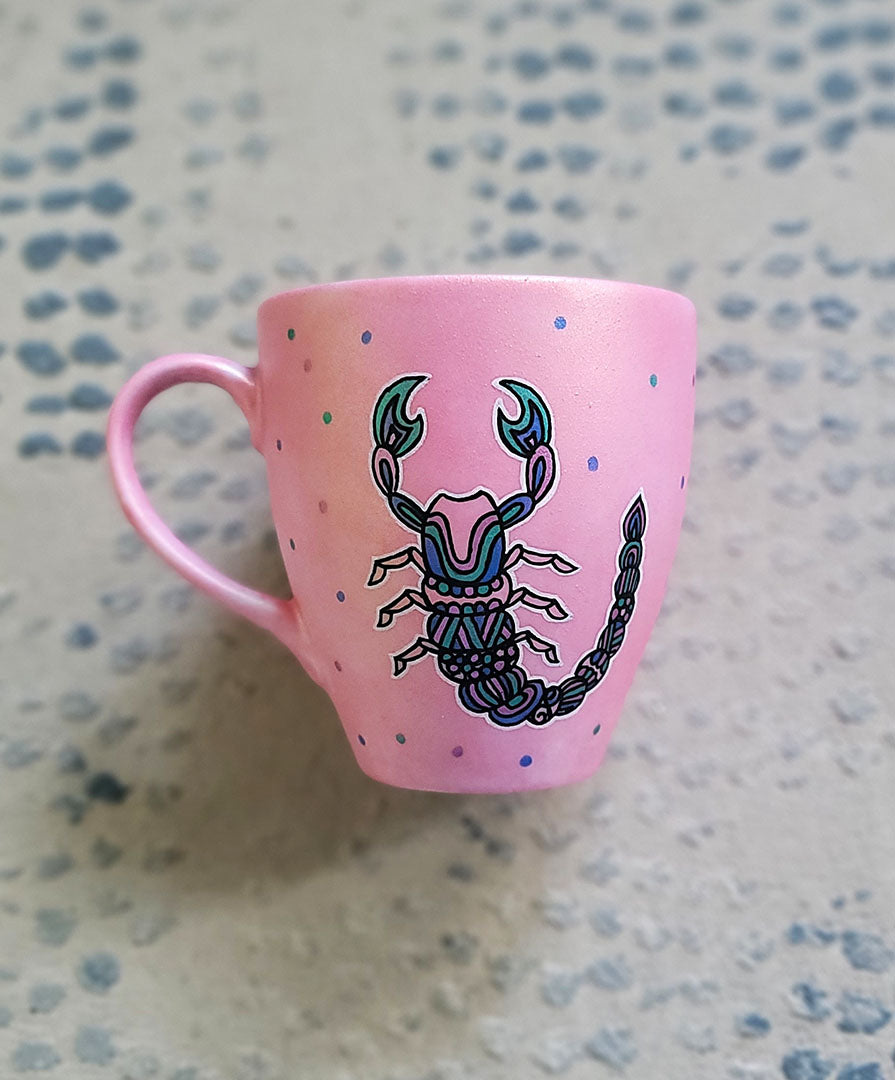 A Shimmery Pink Hand painted Mug With a Scorpio Design on it in Black and colored in pinks, greens and blues and dots scattered all over the mug