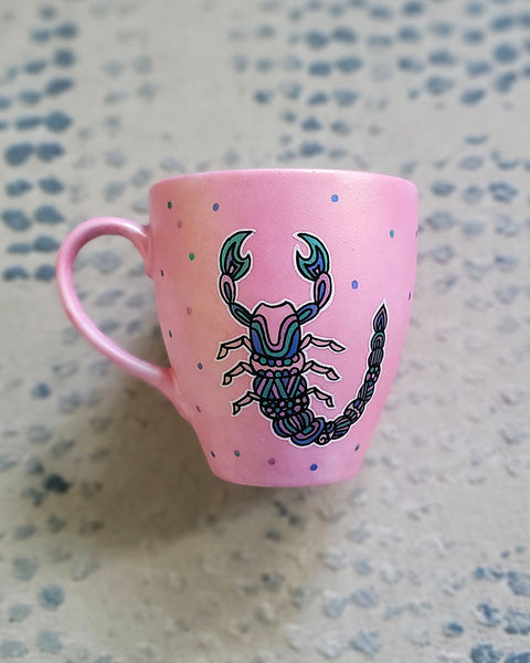 A Shimmery Pink Hand painted Mug With a Scorpio Design on it in Black and colored in pinks, greens and blues and dots scattered all over the mug