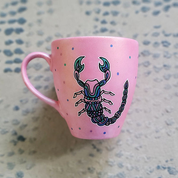 A Shimmery Pink Hand painted Mug With a Scorpio Design on it in Black and colored in pinks, greens and blues and dots scattered all over the mug