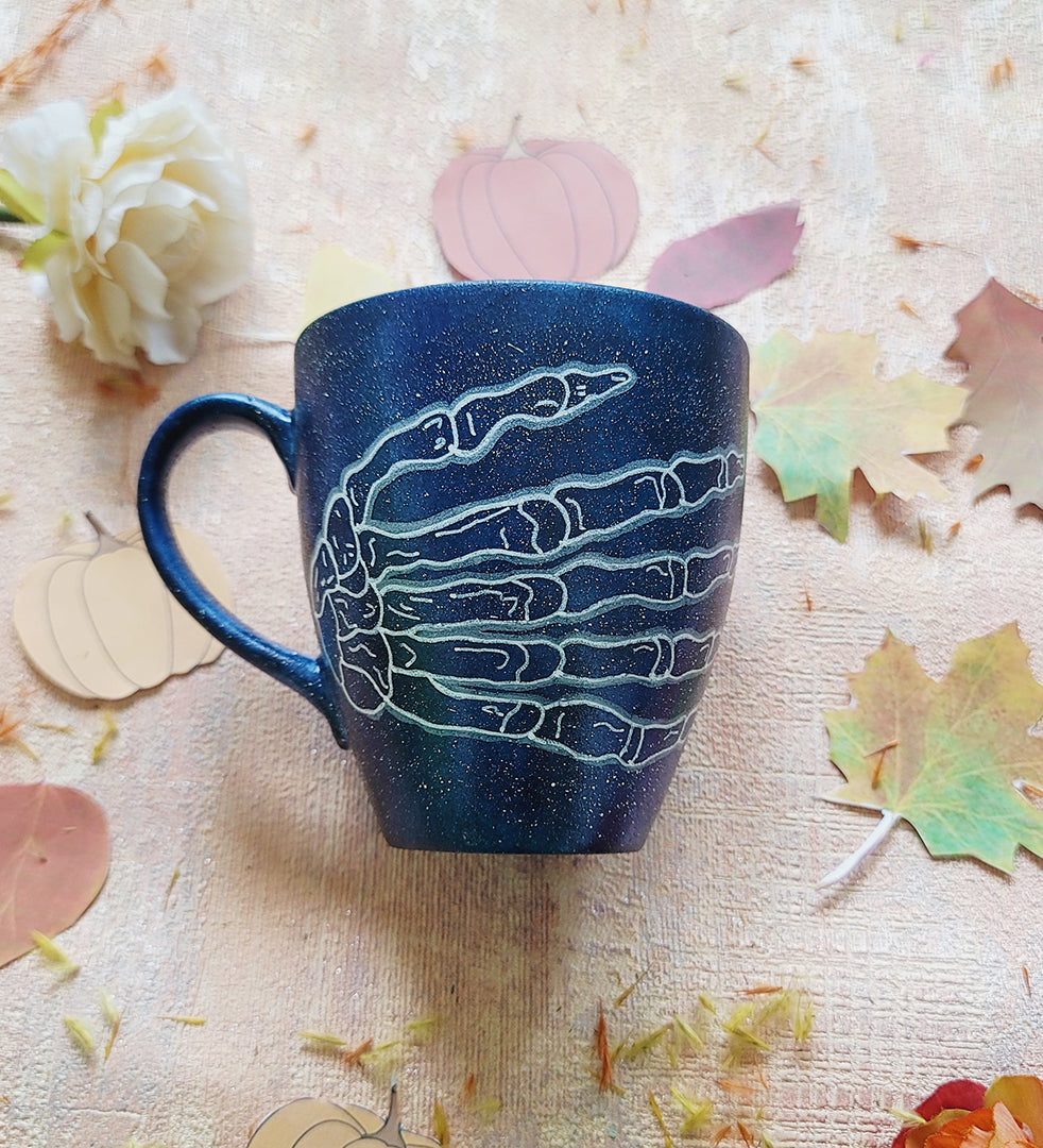 A Bluish Galaxy Hand painted mug with a skeleton hand drawn on it in white