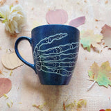 A Bluish Galaxy Hand painted mug with a skeleton hand drawn on it in white