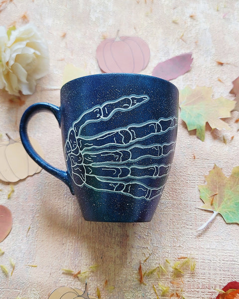 A Bluish Galaxy Hand painted mug with a skeleton hand drawn on it in white