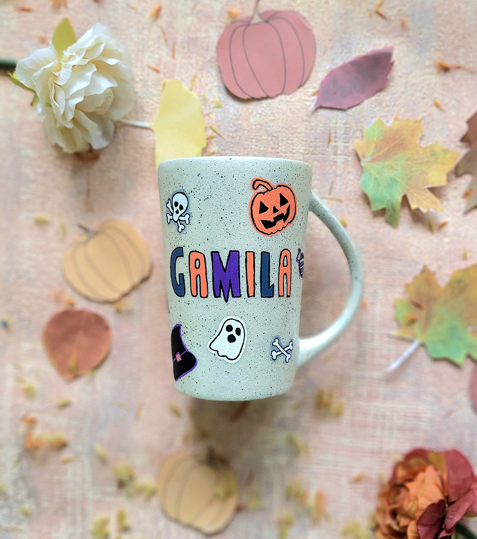 An Ivory hand painted mug with the name "gamila" and Halloween items and black dots scattered all over