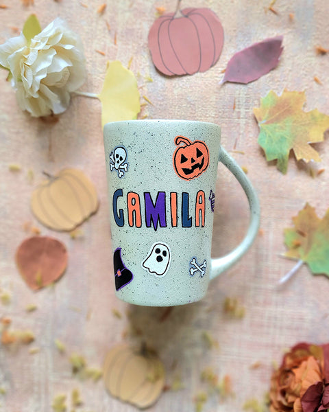 An Ivory hand painted mug with the name "gamila" and Halloween items and black dots scattered all over