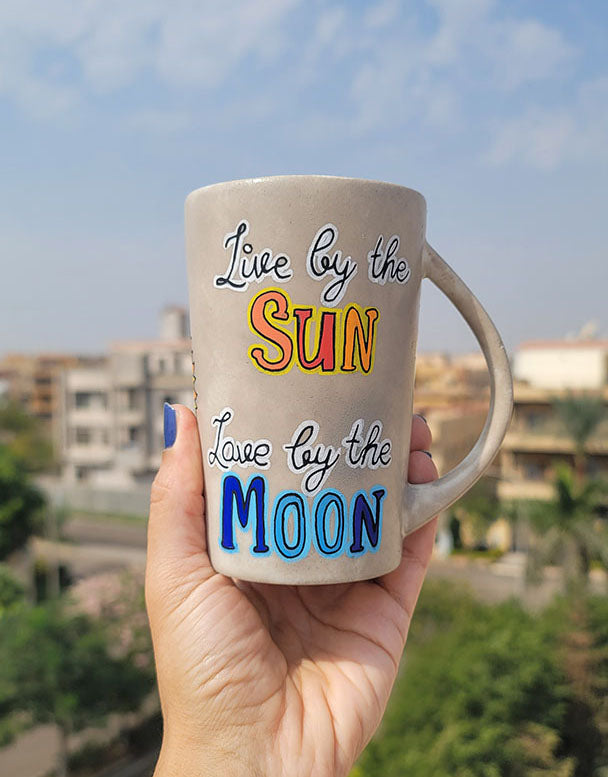 A Taupe hand painted mug with the words live by the (in black and white ) and then the word sun underneath in orange and yellow. then "love by the " written in black and white" and then the word "moon " in blues.