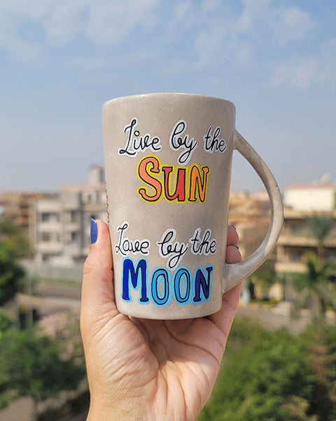 A Taupe hand painted mug with the words live by the (in black and white ) and then the word sun underneath in orange and yellow. then "love by the " written in black and white" and then the word "moon " in blues.