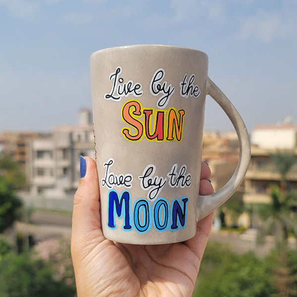 A Taupe hand painted mug with the words live by the (in black and white ) and then the word sun underneath in orange and yellow. then "love by the " written in black and white" and then the word "moon " in blues.