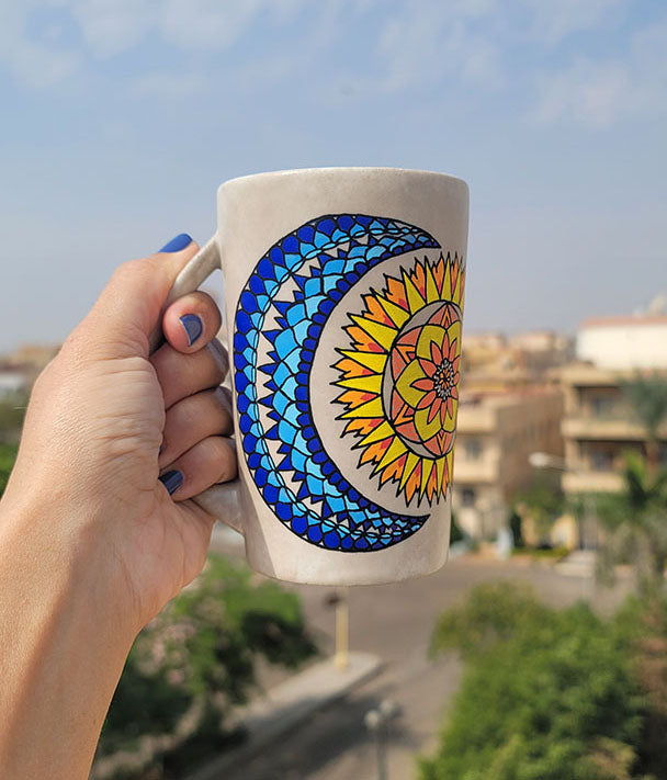 A Taupe hand painted mug with a design of a moon (in colors of blues) and sun ( in colors of yellows and oranges)