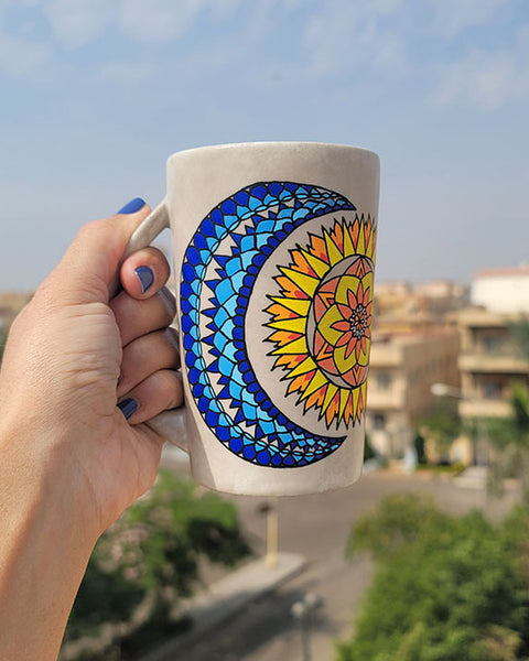 A Taupe hand painted mug with a design of a moon (in colors of blues) and sun ( in colors of yellows and oranges)