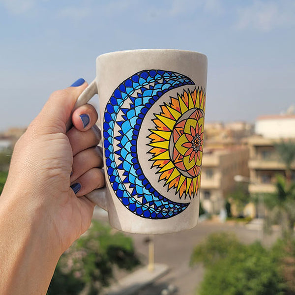 A Taupe hand painted mug with a design of a moon (in colors of blues) and sun ( in colors of yellows and oranges)