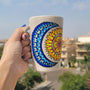 A Taupe hand painted mug with a design of a moon (in colors of blues) and sun ( in colors of yellows and oranges)