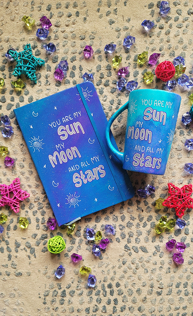 A Blue Galaxy Hand painted mug and notebook with the sentence "you are my Sun, My Moon and all my stars" with small moons stars and suns scattered all over.