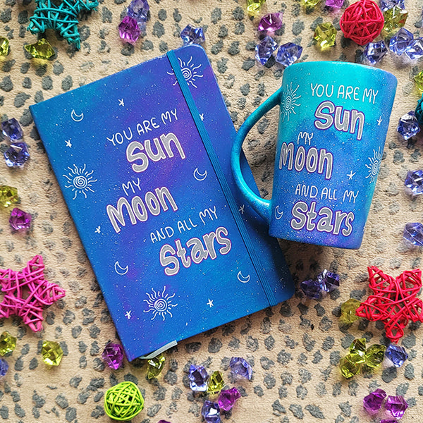 A Blue Galaxy Hand painted mug and notebook with the sentence "you are my Sun, My Moon and all my stars" with small moons stars and suns scattered all over.